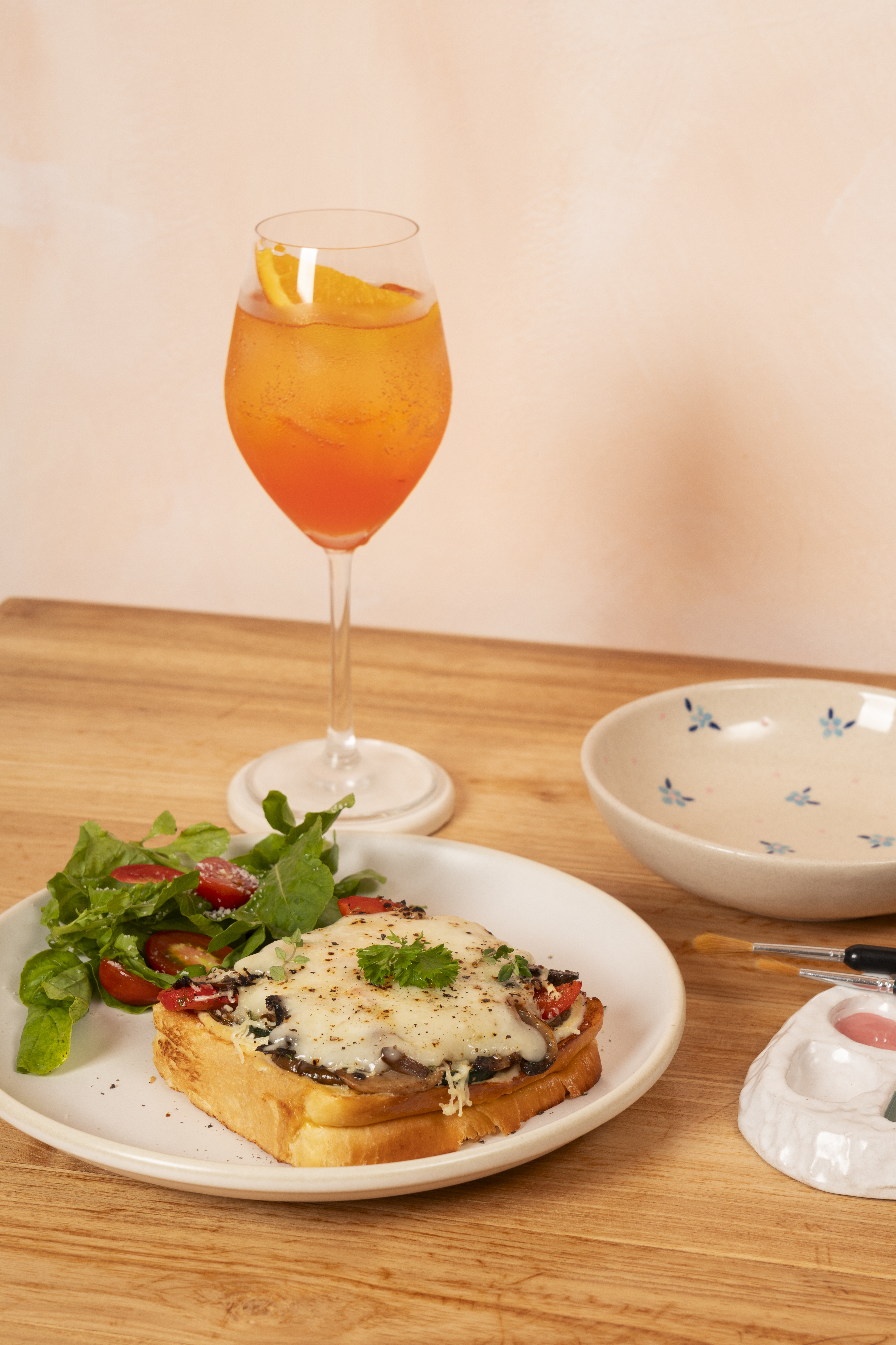 A plated caf&eacute; dish with melted cheese on toast and fresh greens beside a citrus spritz at Klay Bar in Canggu, capturing the relaxed pace of painting ceramics over lunch.